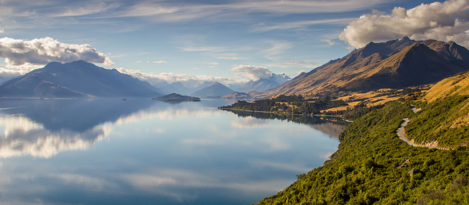 A still mountain lake with snow-dusted peaks and a winding road along the shore